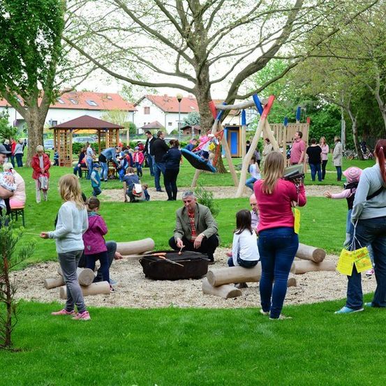 Eine Gruppe von Menschen versammelt sich in einem Park mit einem Feuerkorb, Spielplatzgeräten und Bäumen. Einige sitzen auf Baumstämmen, andere stehen und einige tragen Kinder. Im Hintergrund befinden sich Häuser und ein Pavillon.
