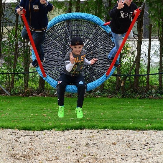 Drei junge Jungen spielen auf einer großen kreisförmigen Schaukel in einem Park, ein Junge sitzt, während die anderen auf den Seiten der Schaukel stehen.