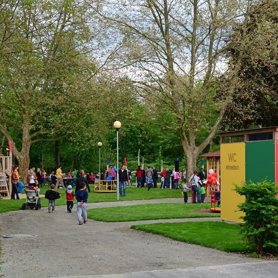 Ein sonniger Tag auf einem Kinderspielplatz mit verschiedenen Spielgeräten, einigen Erwachsenen und Kindern, die herumlaufen, und einem Schild, das auf einen Wintersportbereich hinweist.