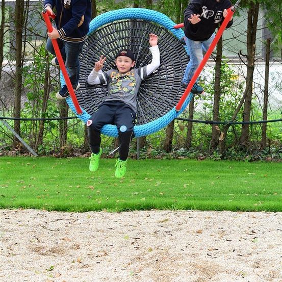 Drei Jungen spielen auf einer großen kreisförmigen Schaukel in einem Park, wobei ein Junge in der Mitte ein Friedenszeichen macht.