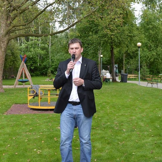 Ein Mann in einem Anzug steht in einem Park, hält ein Mikrofon in der Hand und scheint eine Rede zu halten. Hinter ihm befindet sich ein Spielplatz mit einer gelben Struktur.