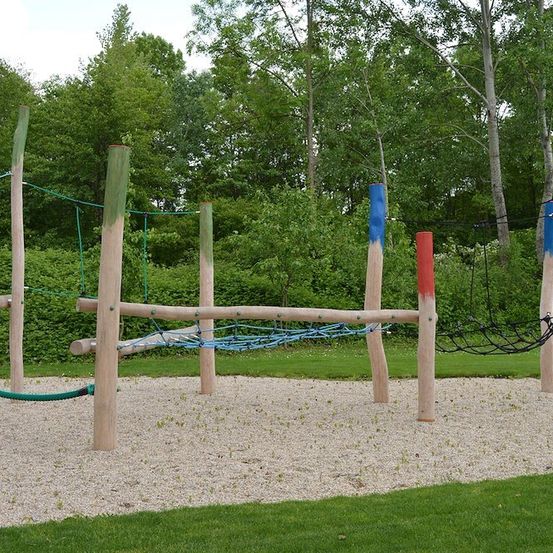 Ein Outdoor-Spielplatz mit Holzpfosten und Netzen, umgeben von grünem Gras und Bäumen.