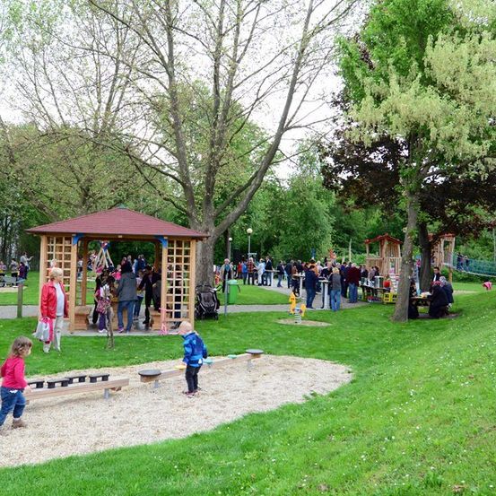 Ein Park mit einem Pavillon, Spielgeräten und Menschen, die spazieren und auf Bänken sitzen. Bäume und Gras umgeben das Gebiet.