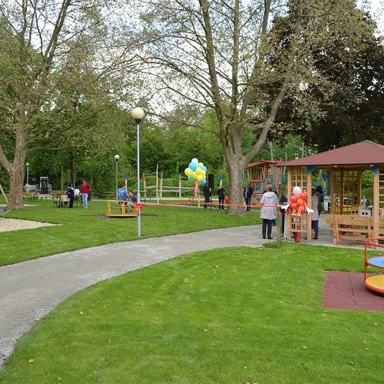 Ein Park mit einem Spielbereich, einem Pavillon und Menschen, die herumlaufen. Es gibt Bäume, Gras und eine rote Bandbarriere.