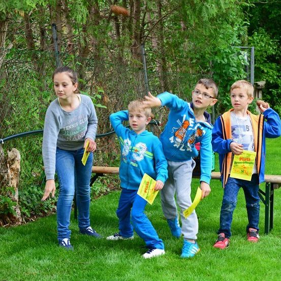 Fünf Kinder stehen auf einem Rasen und halten gelbe Papiere. Ein Junge wirft etwas. Hinter ihnen steht eine Holzbank und ein Zaun.