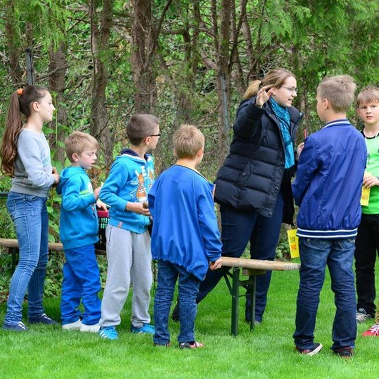 Eine Frau und mehrere Kinder stehen in einem Grasbereich mit einer Holzbank und einem Zaun im Hintergrund. Die Frau spricht zu den Kindern, die sie anschauen.