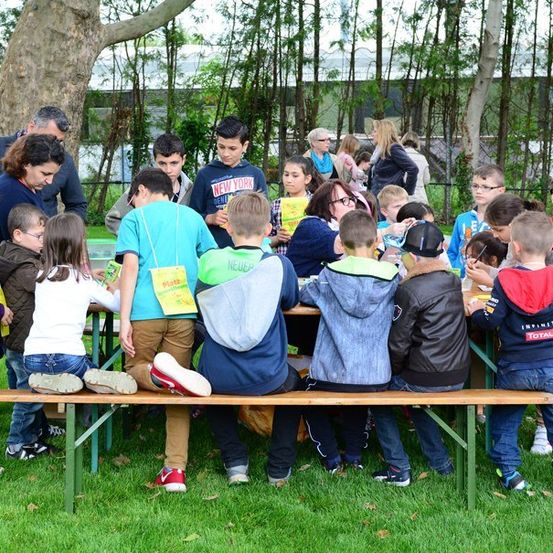Eine Gruppe von Kindern und Erwachsenen versammelt sich um einen Tisch im Park, einige lesen Bücher, während andere in die Kamera schauen.