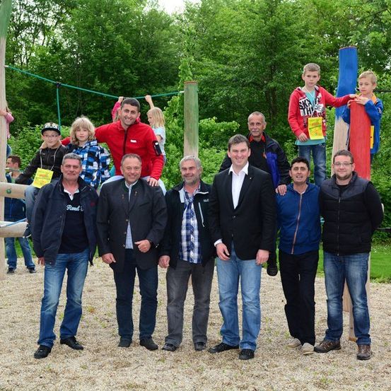 Eine Gruppe von Erwachsenen und Kindern posiert für ein Foto in einem Park mit einer hölzernen Spielstruktur im Hintergrund.
