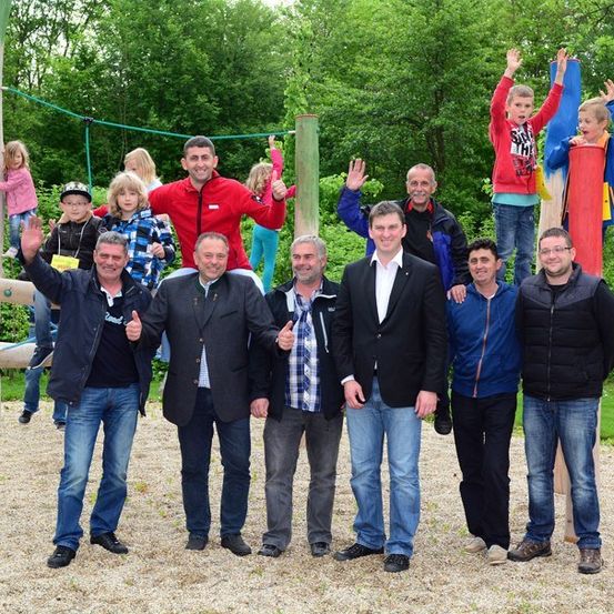 Eine Gruppe von Erwachsenen und Kindern steht zusammen in einem Park, einige auf einer Holzstruktur, mit Bäumen und Gras im Hintergrund.