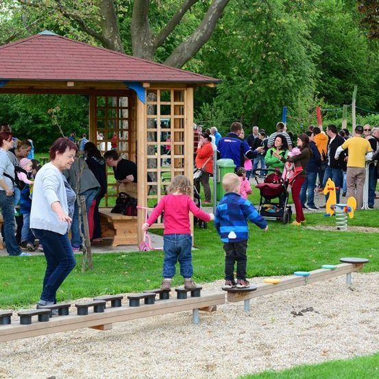 Eine Gruppe von Kindern und Erwachsenen spielt auf einer Wippe in einem Park mit einem Pavillon im Hintergrund.