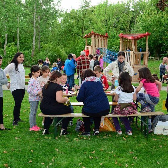 Eine Gruppe von Menschen, einschließlich Erwachsener und Kindern, versammelt sich in einem Park. Einige sitzen auf Bänken, während andere stehen. Eine Frau steht in der Nähe eines Tisches mit einer Box. Im Hintergrund befinden sich Bäume und ein Spielplatz.