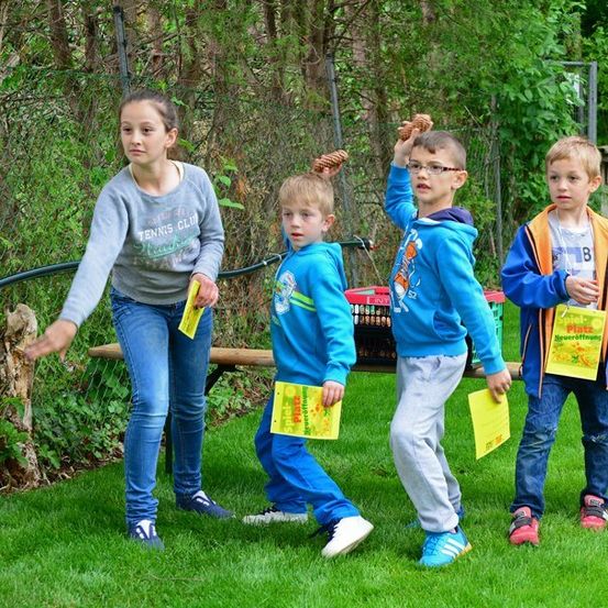 Fünf Kinder stehen auf einem Rasen und halten gelbe Papiere und Tannenzapfen. Sie scheinen an einer Outdoor-Aktivität teilzunehmen. Hinter ihnen steht eine Holzbank und ein Zaun mit Pflanzen.