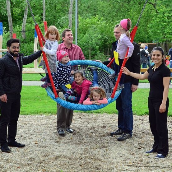 Eine Gruppe von Erwachsenen und Kindern steht um eine blaue Schaukel in einem Park. Die Kinder sitzen in der Schaukel und die Erwachsenen halten die Ketten.