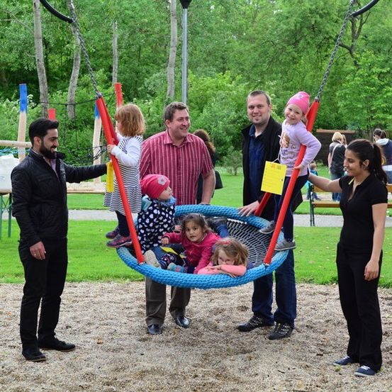 Eine Gruppe von Erwachsenen und Kindern versammelt sich um eine blaue Schaukel in einem Park. Einige Kinder sitzen auf der Schaukel, während andere in der Nähe stehen. Die Erwachsenen lächeln und scheinen für ein Foto zu posieren.