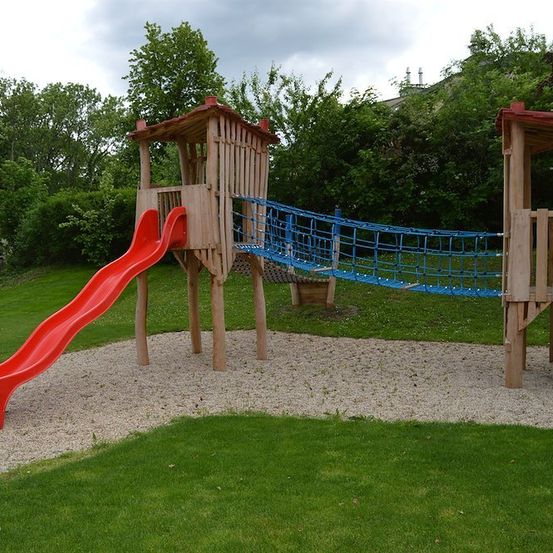 Ein hölzernes Spielgerät mit einer roten Rutsche und einem blauen Kletternetz in einem Rasenbereich.