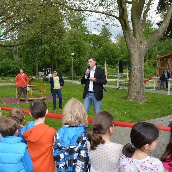 Ein Mann im Anzug spricht in ein Mikrofon in einem Park, während Kinder und Erwachsene zusehen. Im Hintergrund befinden sich Luftballons, ein Baum und Spielplatzgeräte.