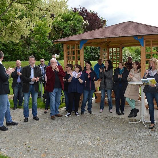 Eine Gruppe von Menschen in formeller Kleidung steht in einem Park, klatscht und lächelt, wobei eine Frau ein Mikrofon und ein Buch hält. Ein hölzernes Pavillon ist im Hintergrund.