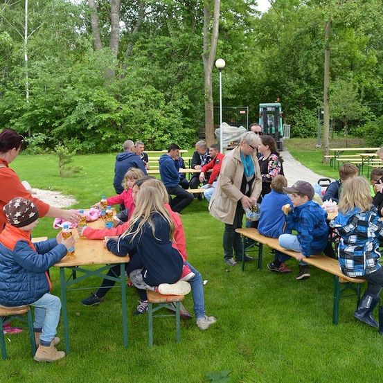 Eine Gruppe von Kindern und Erwachsenen sitzt um Picknicktische in einem Park und isst und trinkt. Der Park ist von Bäumen und Grünflächen umgeben.