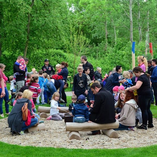 Eine Gruppe von Menschen ist in einem Park versammelt, einige sitzen auf Baumstämmen und andere stehen herum. Ein Mann spielt ein Musikinstrument, während ein junges Mädchen zuschaut.