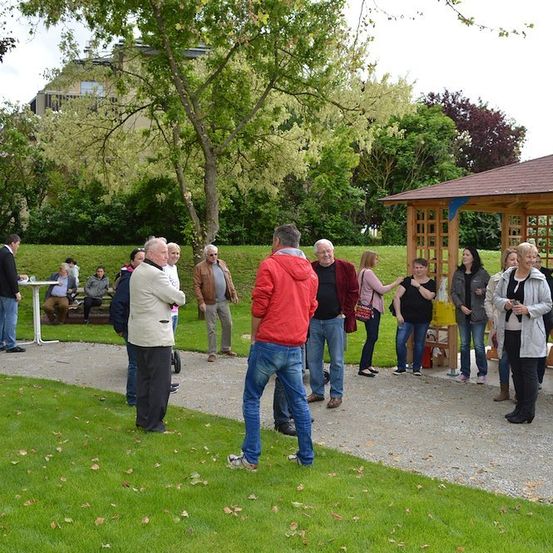 Eine Gruppe von Menschen versammelt sich in einem Park, einige stehen und einige sitzen auf Bänken, mit einem hölzernen Pavillon im Hintergrund.
