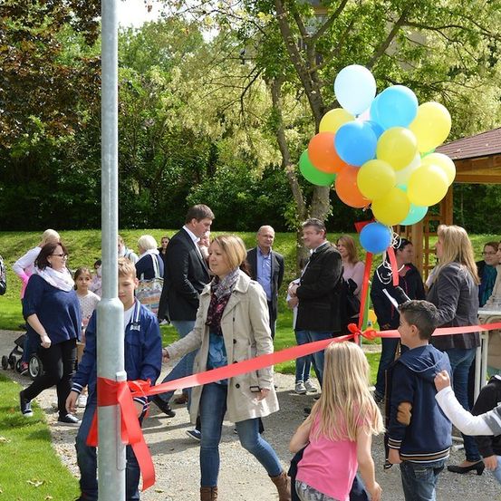 Eine Gruppe von Menschen versammelt sich in einem Park für eine Eröffnungszeremonie. Eine Frau in einem beigen Mantel hält einen Strauß Luftballons. Kinder und Erwachsene sind anwesend, einige halten Gläser.