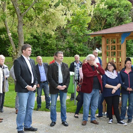 Eine Gruppe von Menschen in lässiger Kleidung steht in einem Park, einige in der Nähe eines hölzernen Pavillons, mit Bäumen und einem Gebäude im Hintergrund.