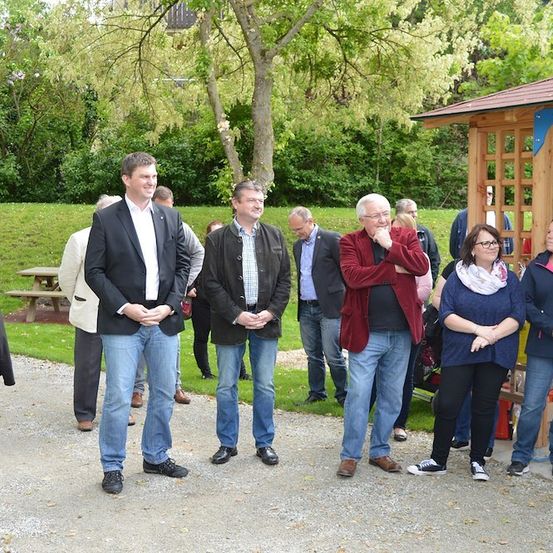 Eine Gruppe von Menschen steht in einem Park, einige lächeln, mit einem hölzernen Pavillon im Hintergrund. Bäume und Grün umgeben sie.