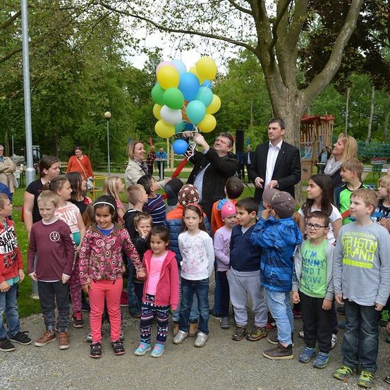 Eine Gruppe von Kindern und Erwachsenen steht in einem Park, ein Mann hält einen Strauß Luftballons. Einige der Kinder tragen Jacken und Turnschuhe. Im Hintergrund befinden sich Bäume, ein Pfosten und ein Spielplatz.