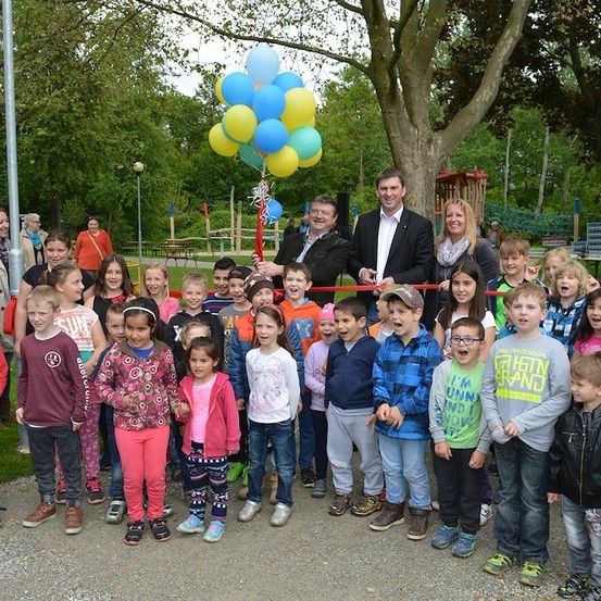 Eine Gruppe von Kindern und Erwachsenen steht in einem Park, lächelt und posiert für ein Foto bei einer Schleifchenschneidezeremonie. Dahinter befindet sich ein Baum mit Luftballons und einem Spielplatz.
