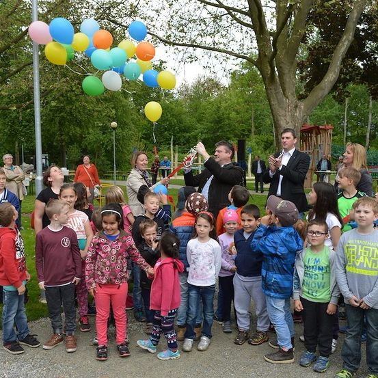 Eine Gruppe von Kindern und Erwachsenen versammelt sich in einem Park mit Luftballons und einem Mann mit Mikrofon.