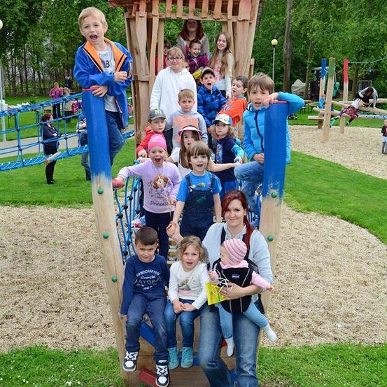 Eine Gruppe von Kindern und Erwachsenen spielt auf einem Spielplatz mit einer Holzstruktur, Kletternetzen und einer Rutsche.