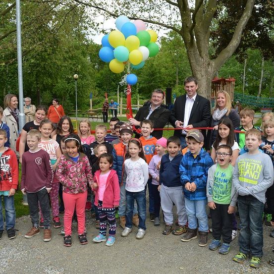 Eine Gruppe von Kindern und Erwachsenen versammelt sich in einem Park und posiert für ein Foto mit Luftballons und einer Schleifenschneidezeremonie.