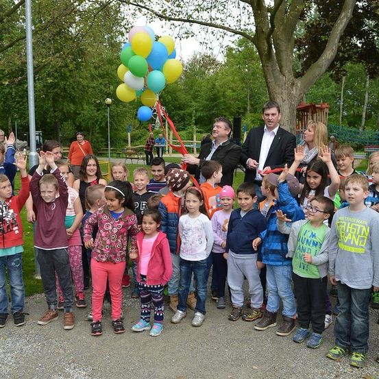 Eine Gruppe von Kindern und Erwachsenen steht in einem Park, einige winken, mit einem Mann, der ein Band vor einem Baum mit Luftballons schneidet.