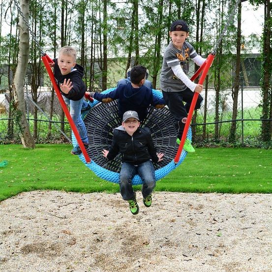 Vier Jungen spielen auf einer kreisförmigen Schaukel in einem Park. Sie tragen Jacken und Turnschuhe. Die Schaukel ist blau und rot. Der Boden ist mit Sand bedeckt. Im Hintergrund befinden sich Bäume und ein Zaun.