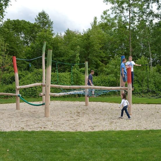 Eine Gruppe von Kindern spielt auf einem hölzernen Hindernisparcours in einem Park mit grünem Gras und Bäumen im Hintergrund.