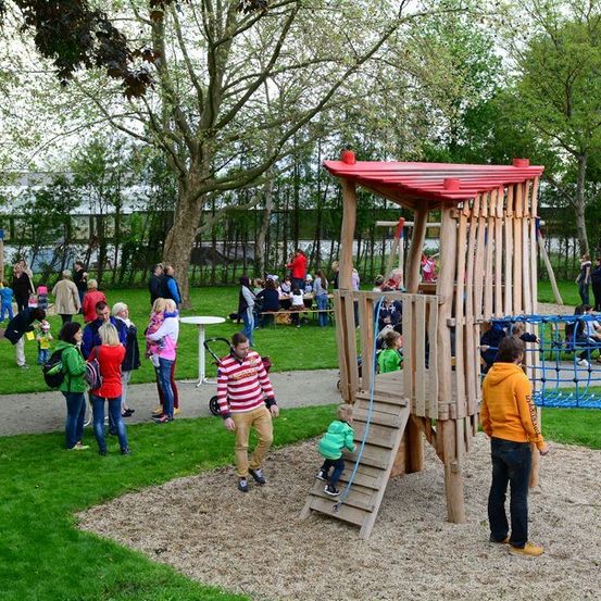 Ein Spielplatz mit einer Holzstruktur, einem roten Dach und einem Kletternetz. Mehrere Menschen gehen herum, einige sitzen auf Bänken und einige spielen auf den Geräten. Der Bereich ist von Bäumen und Gras umgeben.