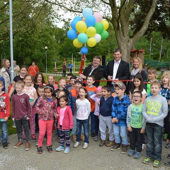 Eine Gruppe von Kindern und Erwachsenen versammelt sich in einem Park, lächelt und posiert für ein Foto mit Luftballons und einer Schleifenschneidezeremonie im Gange.