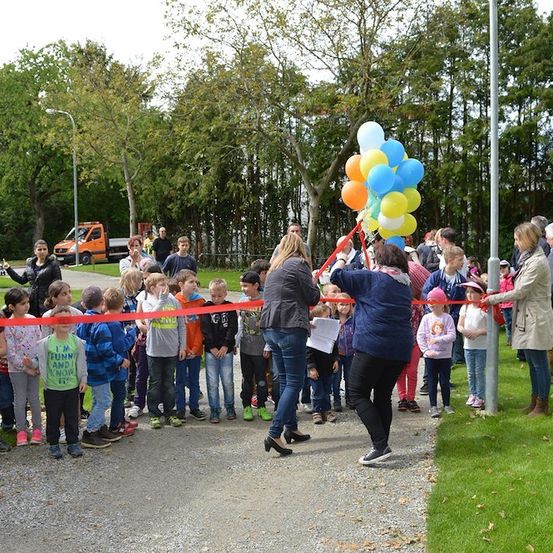 Eine Gruppe von Kindern und Erwachsenen steht um ein Band herum, wobei zwei Frauen Ballons und Scheren zum Schneiden halten. Die Kinder tragen Freizeitkleidung und die Erwachsenen Mäntel. Bäume und ein LKW sind im Hintergrund zu sehen.