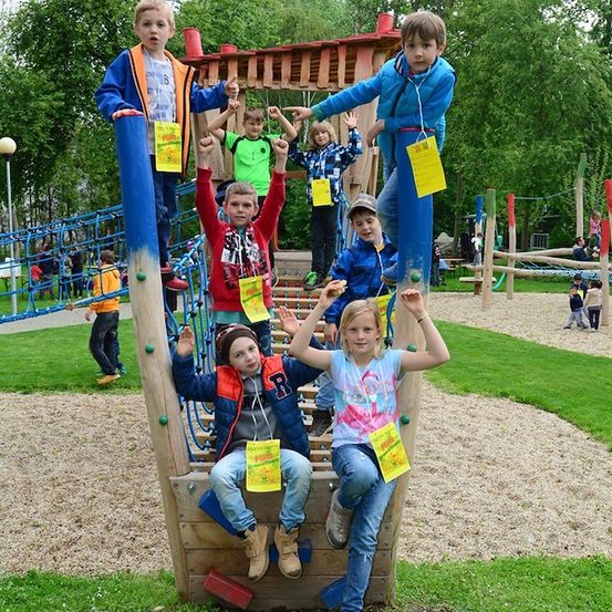 Eine Gruppe von Kindern spielt auf einem Spielplatz mit einem gelben Schild an ihren Shirts. Einige klettern auf die Struktur, während andere auf dem Boden stehen.