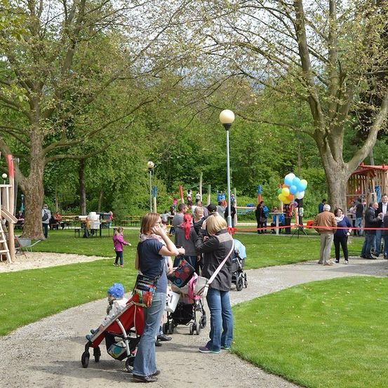 Eine Gruppe von Menschen versammelt sich in einem Park mit einem Spielplatz, Luftballons und einer Banderole. Einige machen Fotos, während andere mit Kinderwagen unterwegs sind.