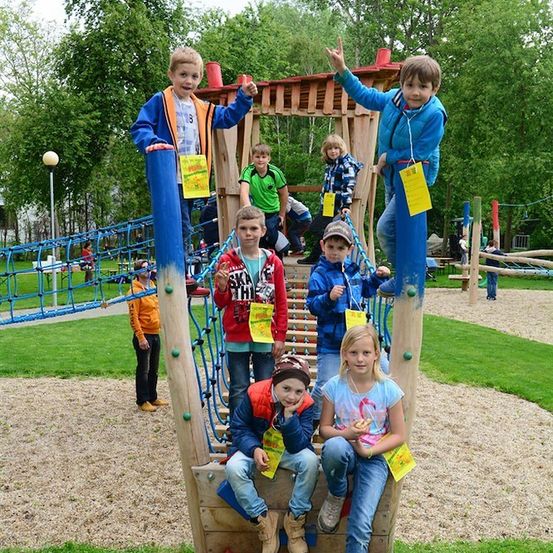 Eine Gruppe von Kindern posiert für ein Foto auf einem Spielplatz, wobei einige auf den Geräten klettern. Sie tragen alle Jacken und halten gelbe Papiere. Der Spielplatz ist von grünem Gras und Bäumen umgeben.