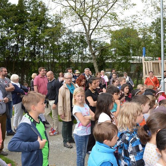 Eine Menschenmenge, bestehend aus Erwachsenen und Kindern, versammelt sich in einem Park, einige schauen sich etwas vor sich an. Das Gebiet ist von Bäumen und einer Holzstruktur umgeben.
