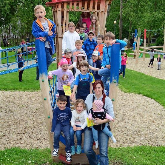 Eine Gruppe von Kindern und Erwachsenen spielt auf einer Holzspielanlage in einem Park. Einige klettern, während andere auf dem Boden sitzen. Die Frau hält ein Baby in einer Trage.