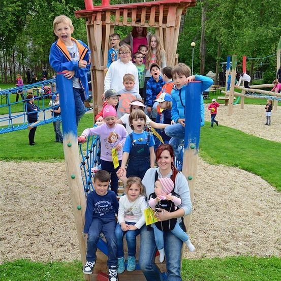 Eine Gruppe von Kindern und Erwachsenen spielt auf einer Holzspielanlage in einem Park. Einige klettern, während andere auf der Anlage sitzen. Eine Frau hält ein Baby in einem Tragetuch.