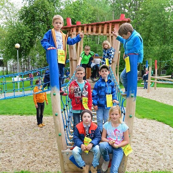 Eine Gruppe von Kindern spielt auf einer hölzernen Spielanlage in einem Park mit grünen Bäumen und Gras. Einige klettern, während andere auf der Anlage sitzen.