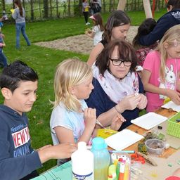Eine Gruppe von Kindern und Erwachsenen ist in einer Outdoor-Kunstaktivität beschäftigt und sitzt um einen Tisch mit verschiedenen Kunstmaterialien. Die Kinder tragen Freizeitkleidung und scheinen die Aktivität zu genießen.