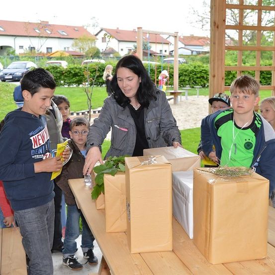Eine Frau verteilt verpackte Geschenke an Kinder in einem Park. Die Kinder lächeln und betrachten die Geschenke.