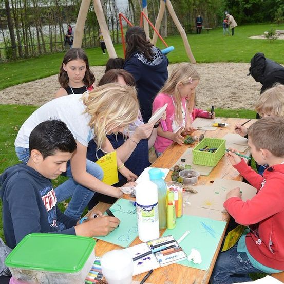 Eine Gruppe von Kindern und Erwachsenen versammelt sich um einen Tisch in einem Park und ist in einer Malaktivität engagiert. Einige benutzen Marker und Farben, während andere Papiere betrachten. Eine Frau führt ein Kind.