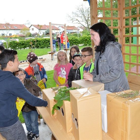 Eine Frau verteilt Pflanzen in Kisten an eine Gruppe von Kindern in einem Park.