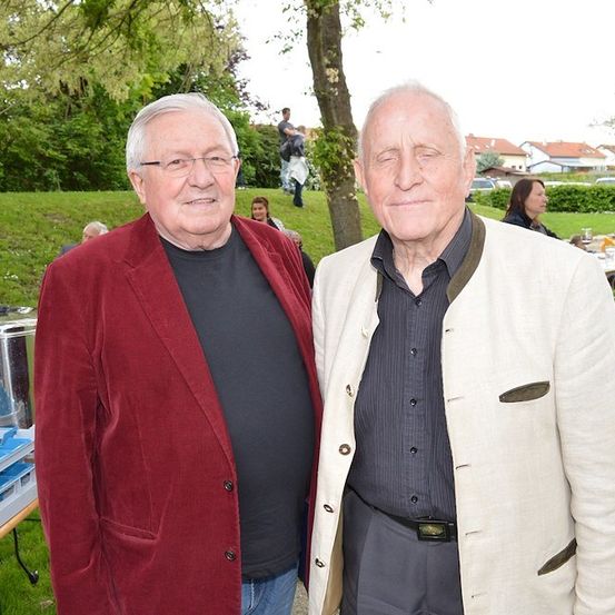 Zwei ältere Männer stehen nebeneinander und lächeln in einem Park mit einem Tisch und Bäumen im Hintergrund.
